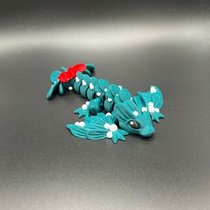 May include: A teal blue and white 3D printed dragon figurine with a red bow on its head. The dragon has a small, round body with wings and a long tail.