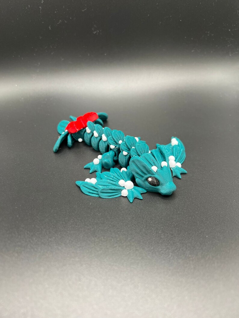 May include: A teal blue and white 3D printed dragon figurine with a red bow on its head. The dragon has a small, round body with wings and a long tail.