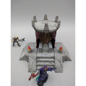May include: A gray stone throne with a red fabric canopy, surrounded by a stone platform with two small torches. The throne is decorated with intricate carvings and is suitable for a miniature game or tabletop role-playing game.
