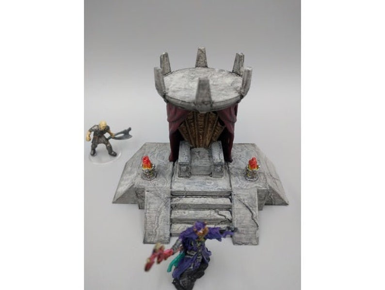May include: A gray stone throne with a red fabric canopy, surrounded by a stone platform with two small torches. The throne is decorated with intricate carvings and is suitable for a miniature game or tabletop role-playing game.