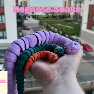 May include: Three 3D printed articulated snake fidget toys. The largest snake is purple, the middle snake is green, and the smallest snake is orange.  The snakes are all in a curled position and have realistic features.  The text "Hognose Snake 3D Printed Articulated Fidget Toy" is at the top of the image.  The text "Fat Girl Makes" is at the bottom of the image.