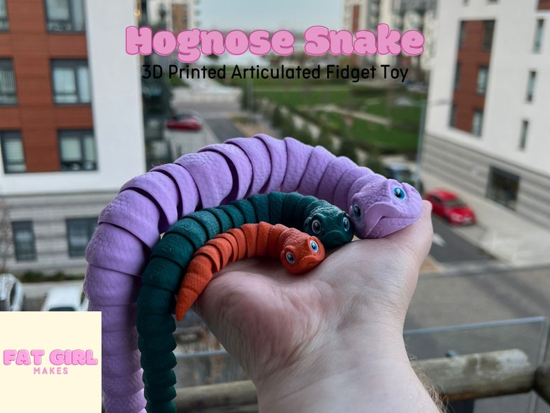 May include: Three 3D printed articulated snake fidget toys. The largest snake is purple, the middle snake is green, and the smallest snake is orange.  The snakes are all in a curled position and have realistic features.  The text "Hognose Snake 3D Printed Articulated Fidget Toy" is at the top of the image.  The text "Fat Girl Makes" is at the bottom of the image.
