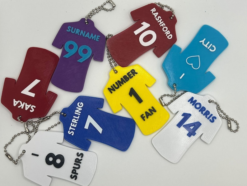 May include: A set of nine colourful plastic jersey-shaped keychains with names and numbers printed on them. The keychains are in various colours, including red, purple, blue, yellow, white, and turquoise.