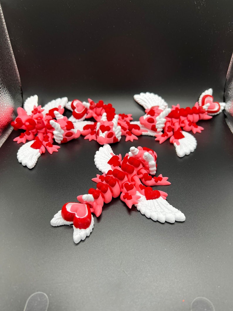 Puede incluir: Cinco criaturas aladas de color rosa y rojo con corazones en sus cuerpos. Las alas son blancas con un borde festoneado. Las criaturas est&aacute;n dispuestas en un grupo sobre un fondo negro.