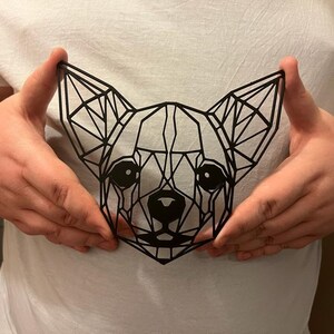 May include: A black geometric design of a chihuahua dog, with a triangular pattern, is held in front of a white t-shirt.