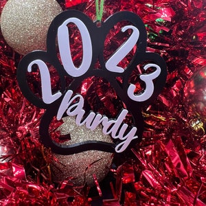 May include: Black paw print Christmas ornament with white 2023 and the name "Rudy" written inside.