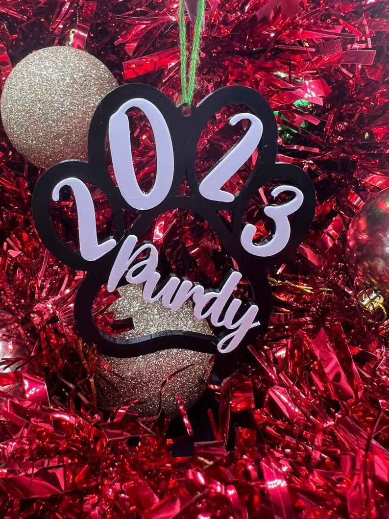 May include: Black paw print Christmas ornament with white 2023 and the name "Rudy" written inside.