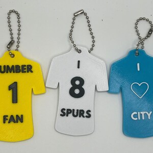 May include: Three keychains shaped like football jerseys. The first keychain is yellow with the number 1 and the text "NUMBER 1 FAN". The second keychain is white with the number 8 and the text "I 8 SPURS". The third keychain is blue with a heart and the text "I CITY".