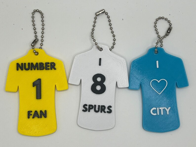 May include: Three keychains shaped like football jerseys. The first keychain is yellow with the number 1 and the text "NUMBER 1 FAN". The second keychain is white with the number 8 and the text "I 8 SPURS". The third keychain is blue with a heart and the text "I CITY".