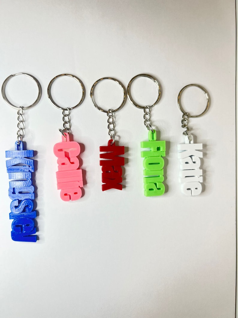 Op de afbeelding: Vijf sleutelhangers met verschillende namen. De namen zijn "Vanessa", "Callie", "Max", "Fiona" en "Katie". De sleutelhangers zijn allemaal zilverkleurig en hebben een ronde ring aan de bovenkant.
