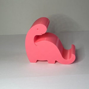 May include: A pink plastic dinosaur phone stand. The dinosaur is in a sitting position with its head tilted back and its tail curled around its body.