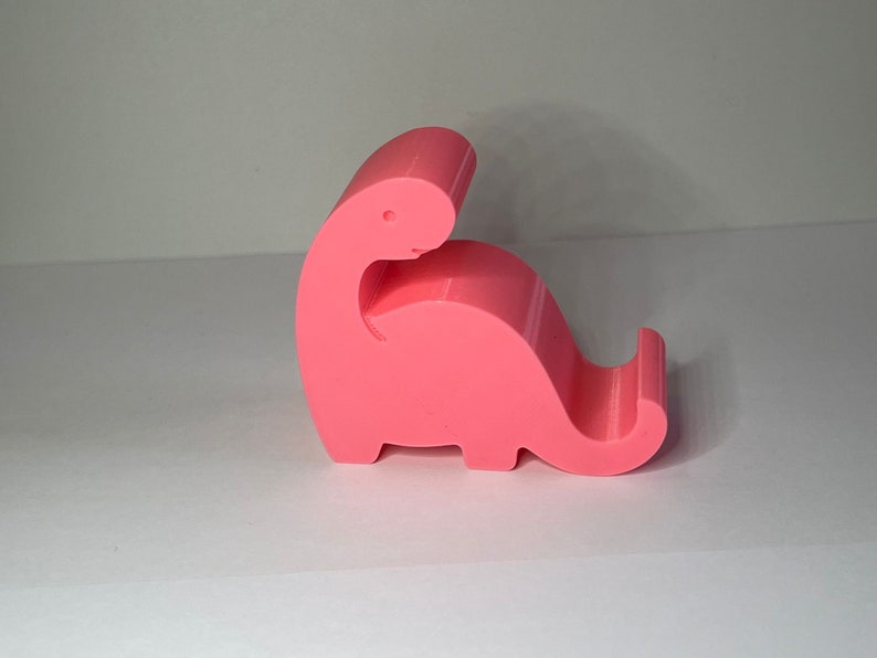May include: A pink plastic dinosaur phone stand. The dinosaur is in a sitting position with its head tilted back and its tail curled around its body.
