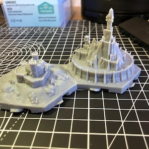 Op de afbeelding: Tweedelig grijs miniatuur kasteelmodel met gedetailleerde torentjes, muren en een bergachtige basis. Het model is gemaakt van een lichtgrijs materiaal en staat op een zwart-wit rasteroppervlak. Het kasteel heeft een middeleeuws thema.