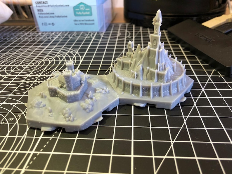 Op de afbeelding: Tweedelig grijs miniatuur kasteelmodel met gedetailleerde torentjes, muren en een bergachtige basis. Het model is gemaakt van een lichtgrijs materiaal en staat op een zwart-wit rasteroppervlak. Het kasteel heeft een middeleeuws thema.