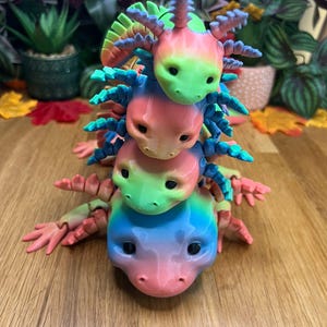 May include: A stack of four colorful axolotl figurines, each with a different rainbow gradient. The axolotls are stacked on top of each other, with the largest axolotl at the bottom and the smallest axolotl at the top.