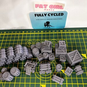 May include: A collection of 3D printed miniature barrels and crates in a gray color. The miniatures are arranged on a green cutting mat with a ruler in the background. The text "FULLY CYCLED" is visible on a blue card in the upper left corner of the image.
