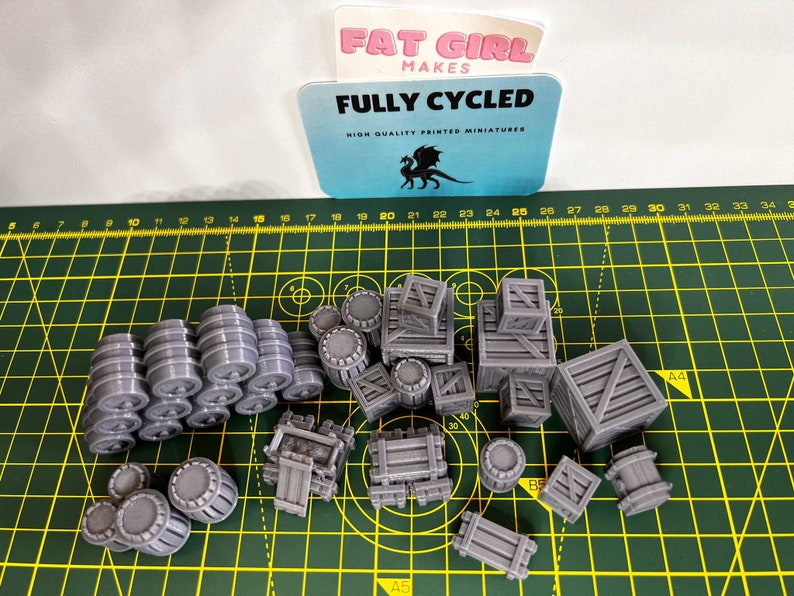 May include: A collection of 3D printed miniature barrels and crates in a gray color. The miniatures are arranged on a green cutting mat with a ruler in the background. The text "FULLY CYCLED" is visible on a blue card in the upper left corner of the image.
