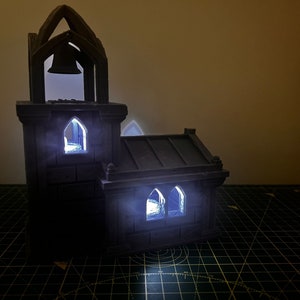 May include: A small, gray, 3D printed model of a church with a bell tower and windows. The windows are lit from inside, creating a warm glow.