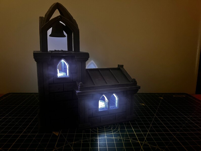 May include: A small, gray, 3D printed model of a church with a bell tower and windows. The windows are lit from inside, creating a warm glow.