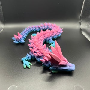 May include: A 3D printed, pink and blue, crystal-like lizard figurine. The lizard is curled up in a serpentine pose with its head facing the right side of the image.