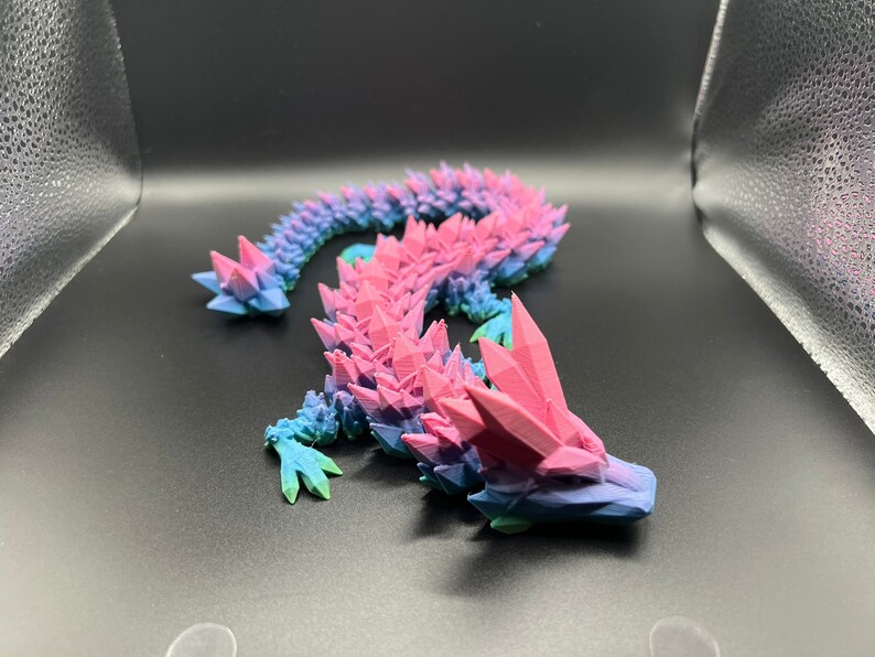 May include: A 3D printed, pink and blue, crystal-like lizard figurine. The lizard is curled up in a serpentine pose with its head facing the right side of the image.