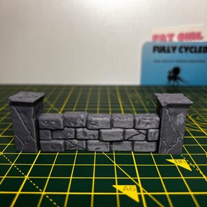 May include: A miniature stone wall with two pillars, made of gray plastic. The wall is made of small, rectangular bricks with a rough texture. The pillars are tall and slender, with a slightly rounded top. The wall is sitting on a green cutting mat.
