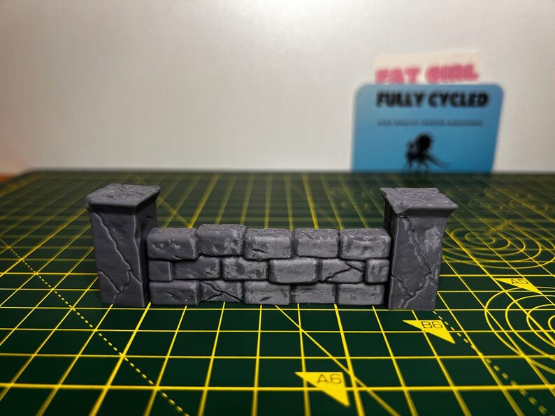 May include: A miniature stone wall with two pillars, made of gray plastic. The wall is made of small, rectangular bricks with a rough texture. The pillars are tall and slender, with a slightly rounded top. The wall is sitting on a green cutting mat.