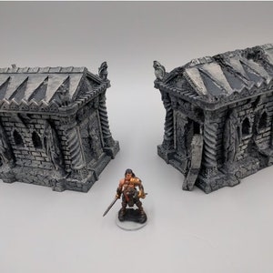 May include: Two gray stone miniature buildings with intricate details, resembling ancient tombs or mausoleums. The buildings have a weathered, aged appearance and feature arched doorways and windows. A miniature figure of a warrior stands in front of the buildings.