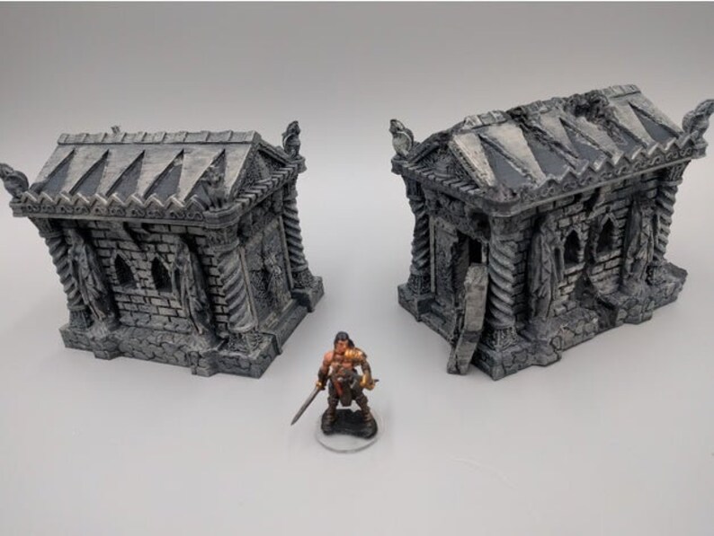 May include: Two gray stone miniature buildings with intricate details, resembling ancient tombs or mausoleums. The buildings have a weathered, aged appearance and feature arched doorways and windows. A miniature figure of a warrior stands in front of the buildings.