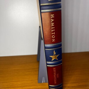 May include: A blue and red hardcover book with gold lettering. The title is "Hamilton: The Revolution". The book is by Melcher Media.