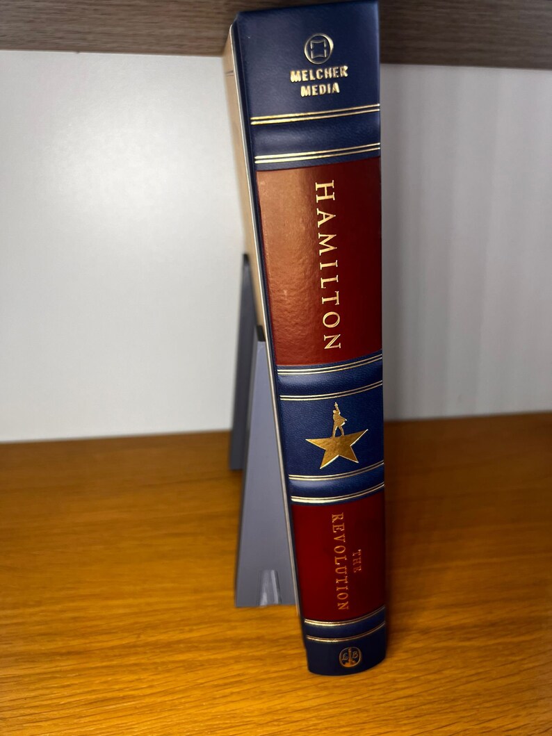 May include: A blue and red hardcover book with gold lettering. The title is "Hamilton: The Revolution". The book is by Melcher Media.