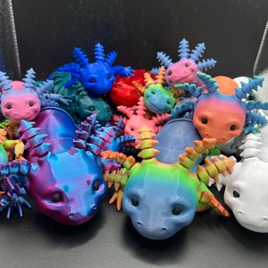 May include: A collection of colorful, 3D printed axolotl figurines. The axolotls are in various poses and have detailed features, including eyes, gills, and fins. The figurines are made in a variety of colors, including blue, pink, red, green, and white.