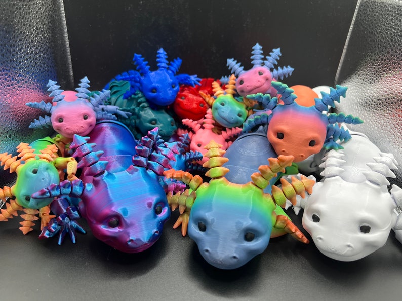 May include: A collection of colorful, 3D printed axolotl figurines. The axolotls are in various poses and have detailed features, including eyes, gills, and fins. The figurines are made in a variety of colors, including blue, pink, red, green, and white.
