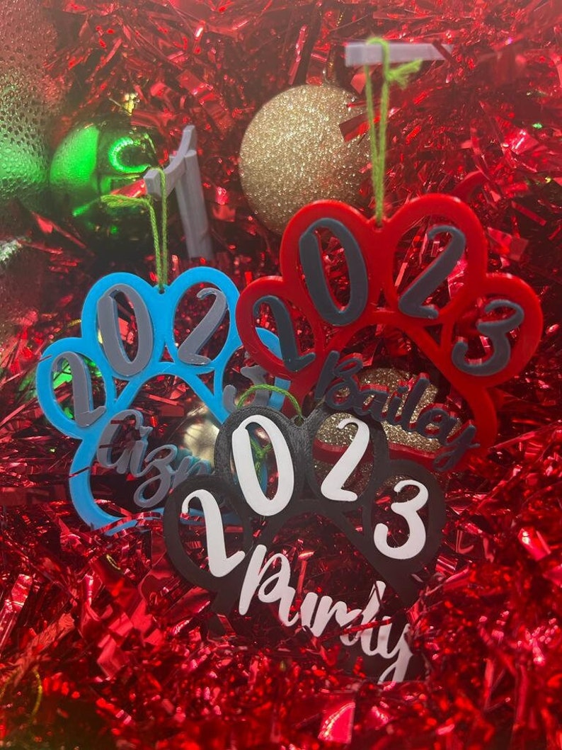 May include: Three paw print shaped Christmas ornaments with the year 2023 and the names "Izzy", "Bailey", and "Pumkin" written on them. The ornaments are blue, red, and black.