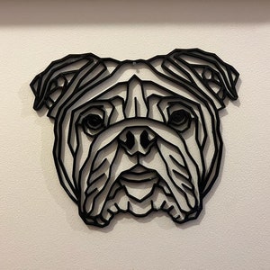 May include: Black geometric wall art of a bulldog's face. The artwork is made of layered lines, creating a 3D effect. The dog's features, including eyes, nose, and jowls, are clearly defined. The art is mounted on a white wall.