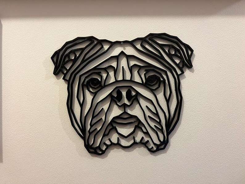 May include: Black geometric wall art of a bulldog's face. The artwork is made of layered lines, creating a 3D effect. The dog's features, including eyes, nose, and jowls, are clearly defined. The art is mounted on a white wall.
