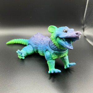 May include: A small, green, blue, and purple plastic toy animal with a toothy grin. The toy is shaped like a small mammal with a long tail and short legs.