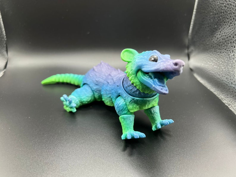 May include: A small, green, blue, and purple plastic toy animal with a toothy grin. The toy is shaped like a small mammal with a long tail and short legs.
