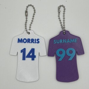 May include: Two white and purple jersey-shaped keychains with blue numbers and names. The white keychain has the name "MORRIS" and the number "14". The purple keychain has the name "SURNAME" and the number "99".