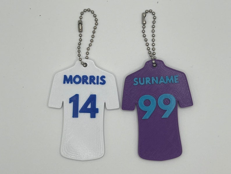 May include: Two white and purple jersey-shaped keychains with blue numbers and names. The white keychain has the name "MORRIS" and the number "14". The purple keychain has the name "SURNAME" and the number "99".