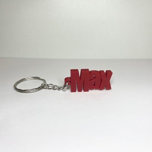 Op de afbeelding: Rode plastic sleutelhanger met de naam "Max" gespeld in blokletters. De sleutelhanger is bevestigd aan een zilveren metalen ring en ketting.