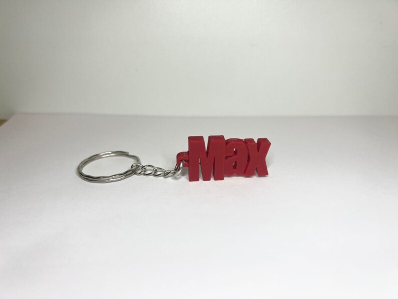 Op de afbeelding: Rode plastic sleutelhanger met de naam "Max" gespeld in blokletters. De sleutelhanger is bevestigd aan een zilveren metalen ring en ketting.