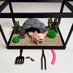 Puede incluir: Un hábitat de ajolote en miniatura con un marco negro, con una base de color arena, una cueva de roca gris y decoraciones de plantas verdes. Un pequeño ajolote de color claro con branquias rosas está en el hábitat. Los accesorios incluyen una pala, pinzas y un gusano rosa.