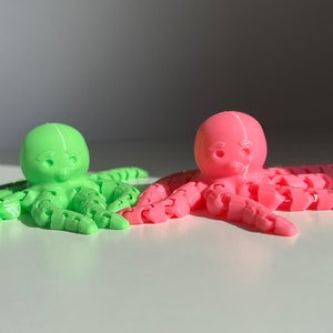 以下が含まれることがあります： 3Dプリントされたタコのおもちゃ2つ。1つは緑色、もう1つはピンク色で、腕はセグメント化されており、かわいい顔をしています。