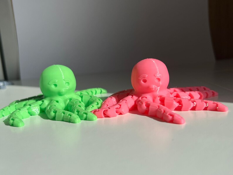 以下が含まれることがあります： 3Dプリントされたタコのおもちゃ2つ。1つは緑色、もう1つはピンク色で、腕はセグメント化されており、かわいい顔をしています。