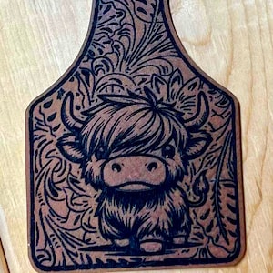Può includere: Un'etichetta in legno marrone con un'illustrazione in bianco e nero di una mucca delle Highlands con un motivo floreale intorno.
