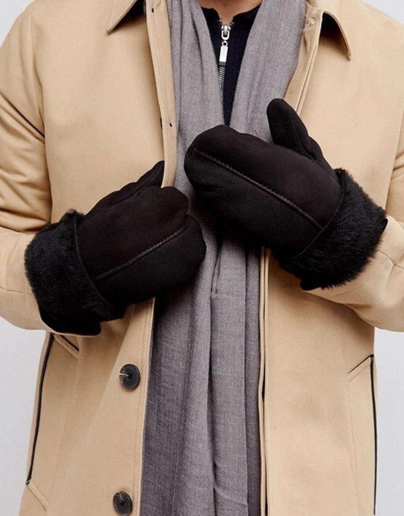 May include: A pair of black sheepskin mittens with a faux fur cuff. The mittens are shown being worn by a person wearing a tan coat.