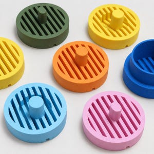 May include: A collection of seven circular, matte-finish objects in various colours including olive green, yellow, orange, light blue, pink, and dark blue. Each piece features a central cylindrical handle and a series of parallel, linear cutouts.