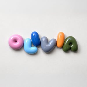 May include: A set of six smooth, glossy, rounded ceramic letters spelling the name OLIVIA. The letters are coloured in pink, light blue, dark blue, grey, orange, and olive green, each featuring a distinct, puffed, and inflated appearance.
