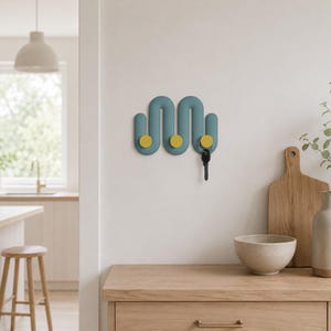 Peut inclure: Un porte-manteau mural bleu sarcelle en forme de vague avec trois patères jaunes arrondies. Le design présente une forme serpentine continue avec des extensions verticales. Un trousseau de clés en métal est suspendu à la patère jaune de droite.